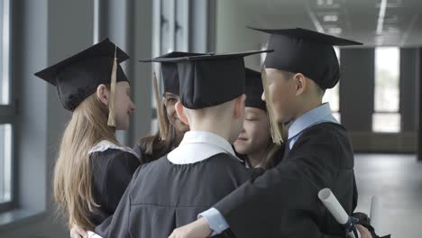 group of happy multiracial preschool students in mortarboard and gown. they are hugging.