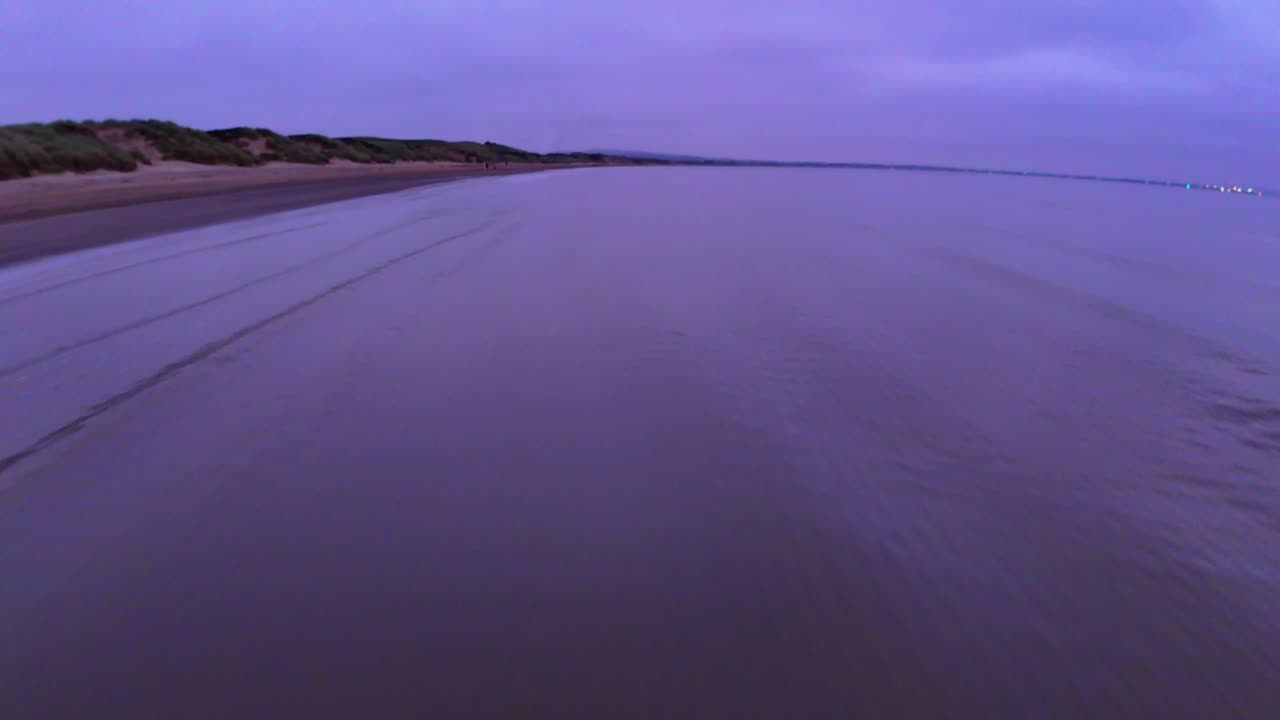 Dynamic low drone flight skimming above a sandy shoreline at sunset, capturing the gentle tide rolling onto the coast with smooth cinematic motion and evening reflections