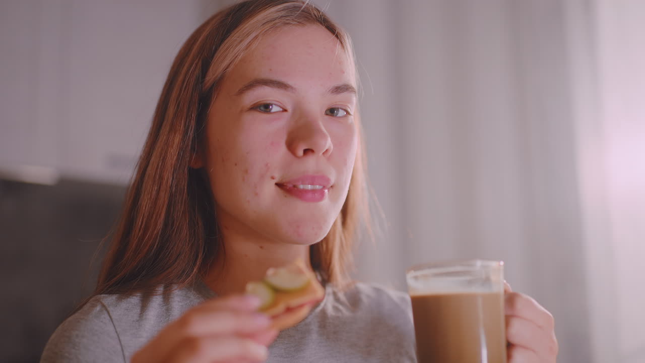 Young lady sipping tea from glass cup while holding sandwich in other hand, soft indoor light reflecting on face and drink, capturing relaxed everyday lifestyle moment of nourishment