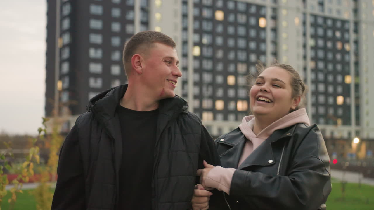 Funny moment between lovers as they take evening stroll in city street she playfully tickles him while laughing he turns to look at her smiling with tall residential buildings glowing behind them