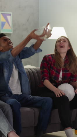 playful black man pours chips onto friends celebrating final goal at football match in sitting room slow motion. joyful soccer fans support players