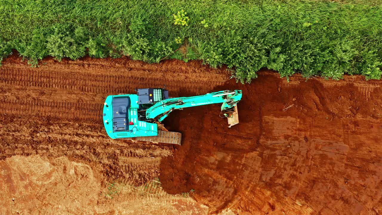 Aerial view of bright blue excavator leveling red soil near green overgrowth