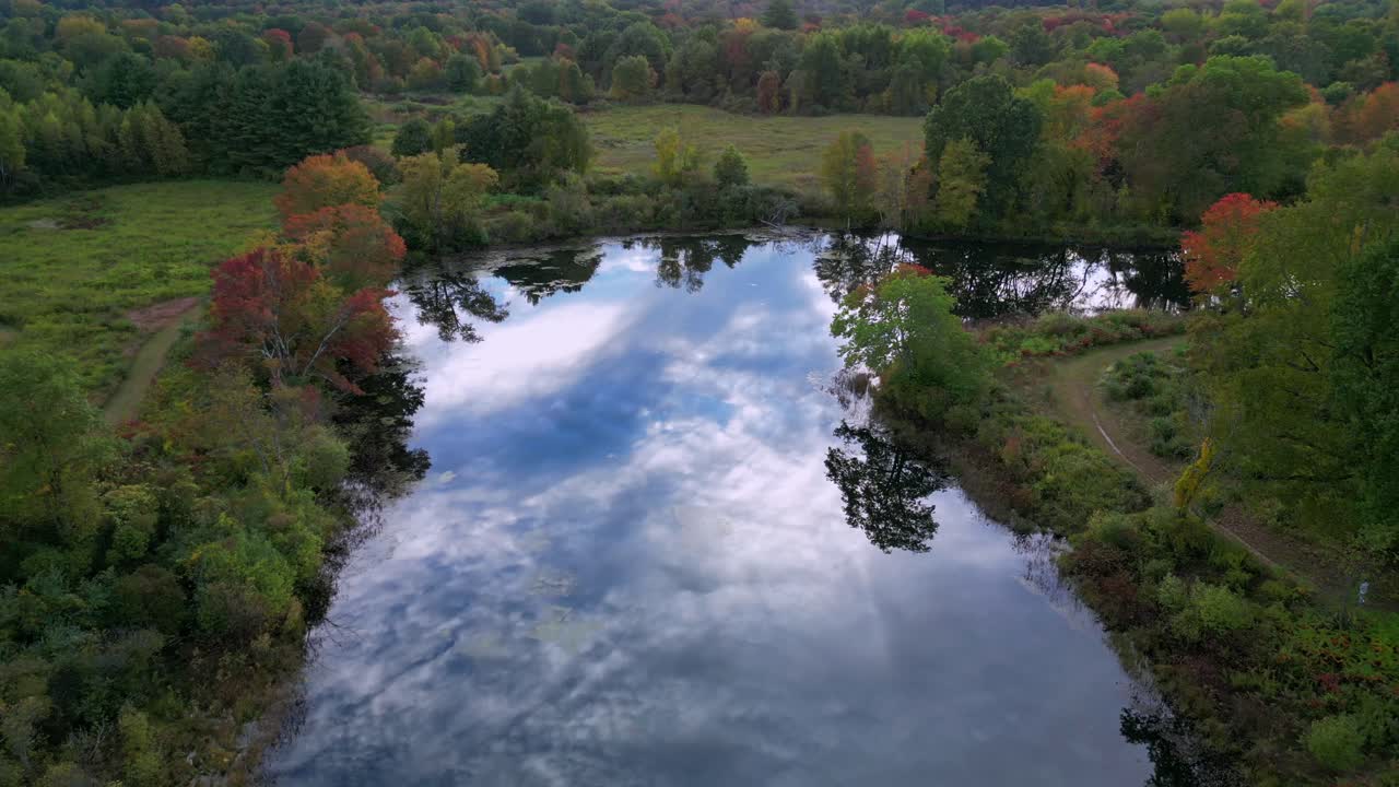 Aerial panoramic dolly above pond with calm water reflecting textured sky and clouds in middle of green forest