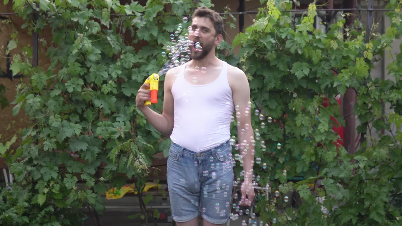 수염을 가진 어린 소년이 서서 장난감 거품에서 비누 거품을 던지고 입으로 잡으려고 합니다.