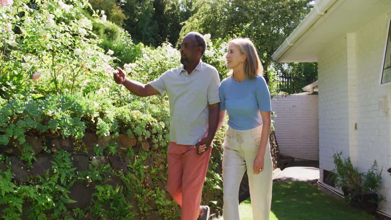 una pareja mayor diversa caminando en su jardín al sol tomándose de las manos y hablando