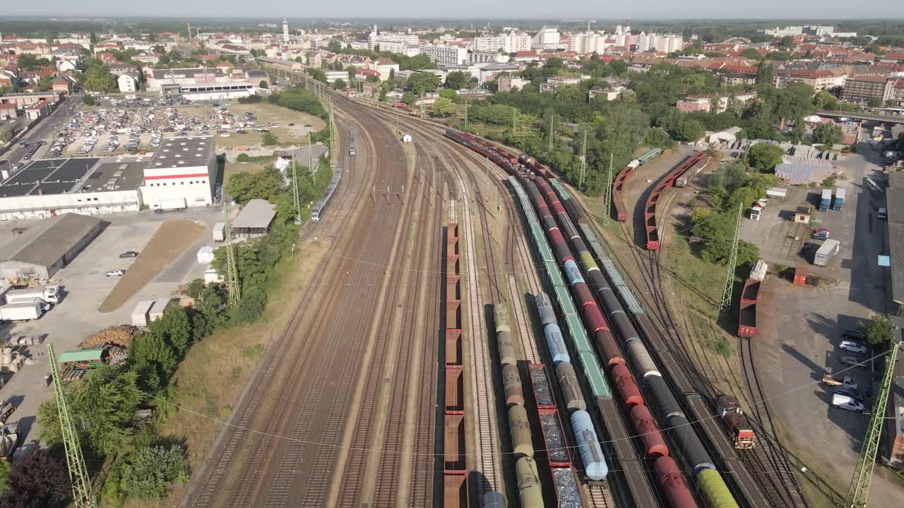 An aerial video shows multiple freight trains stationed in a busy railyard on the edge of a European city. The drone flying over of colorful cargo wagons.