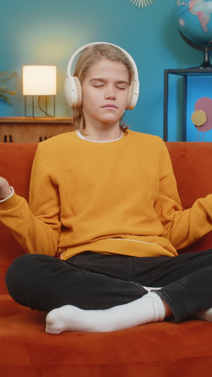 Teenager boy relaxing on home sofa meditating in lotus pose with headphones after long school day