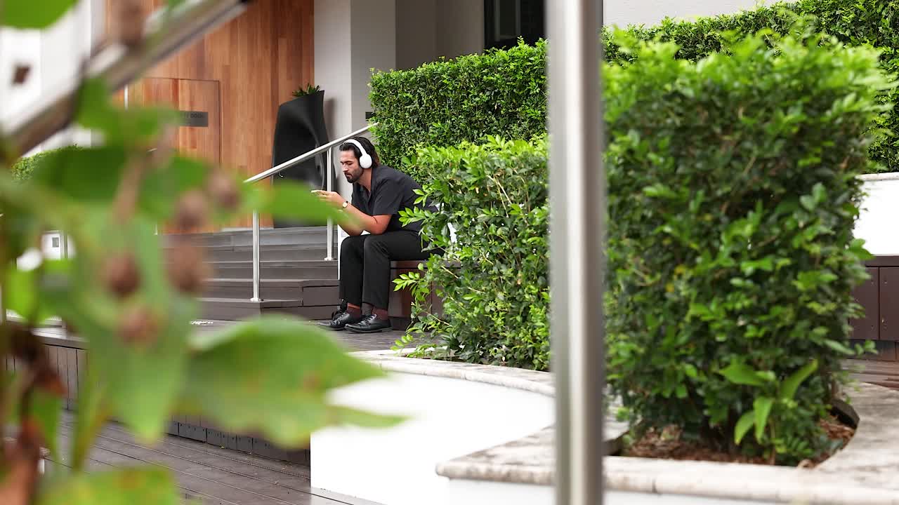 A man sits on steps, listening to music with headphones, surrounded by lush greenery in a sunny urban setting