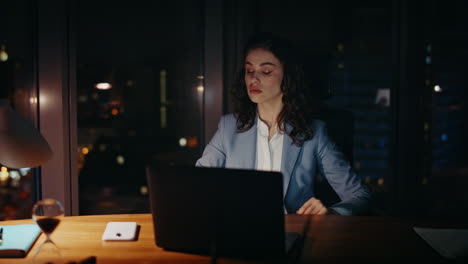 mujer de negocios sobrecargada buscando portátil sentada en el escritorio de la oficina tarde en la noche primer plano
