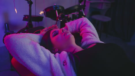 elegant woman lies back in gaming chair with hands on head, bathed in moody neon light, drum set close behind her, surrounded by creative studio equipment in ambient relaxed setting