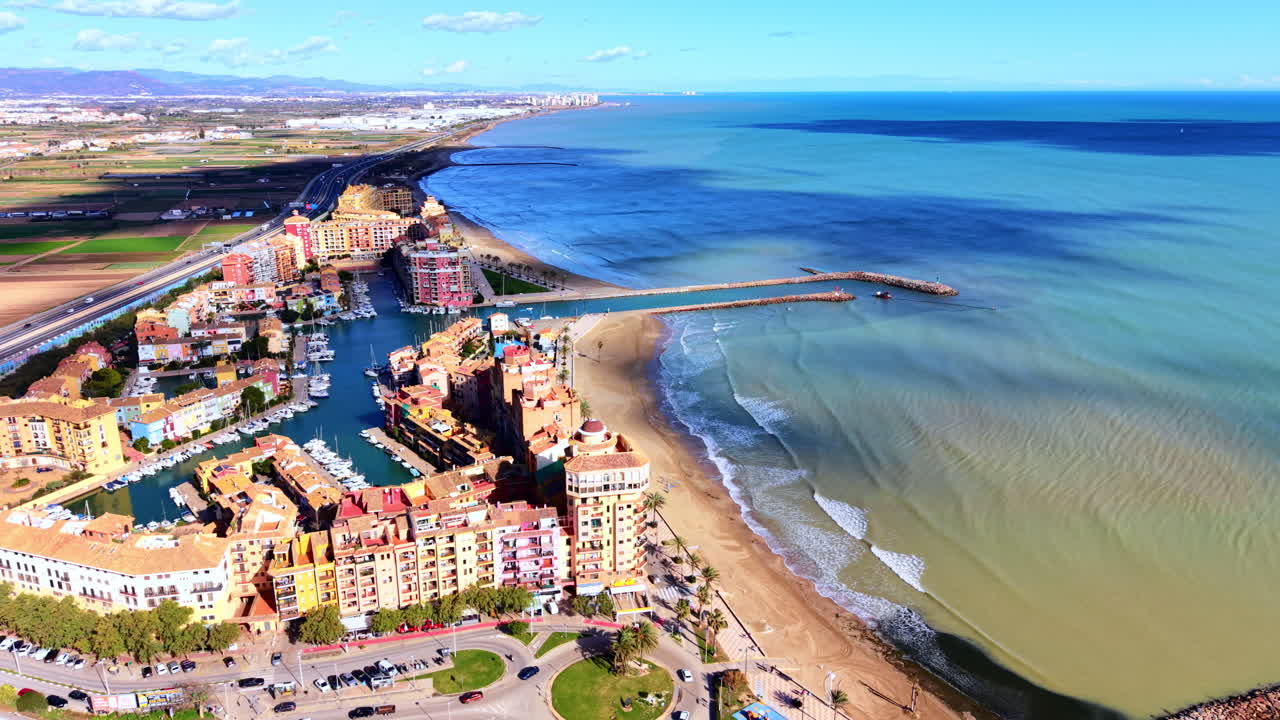 Panorama over en havneby med farverige lejligheder, hvide yachter, strand og en motorvej, der løber langsmed. Højvinkel luftfoto af et luksuriøst kystresort i Spanien.