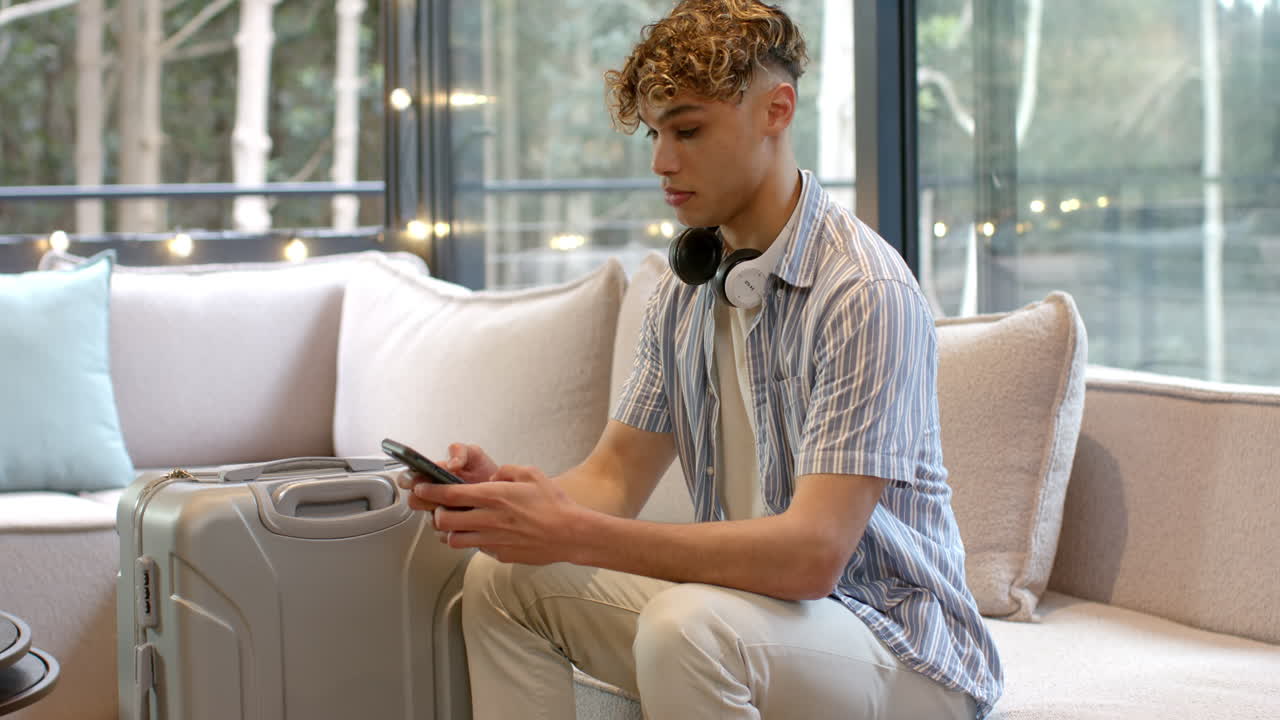 Young man with headphones using smartphone, sitting with suitcase at home