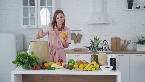 pretty woman taking out fresh fruit from paper shopping bag at home. young female holds an orange in the light kitchen. healthy fruit and vegetables on the table. vertical video