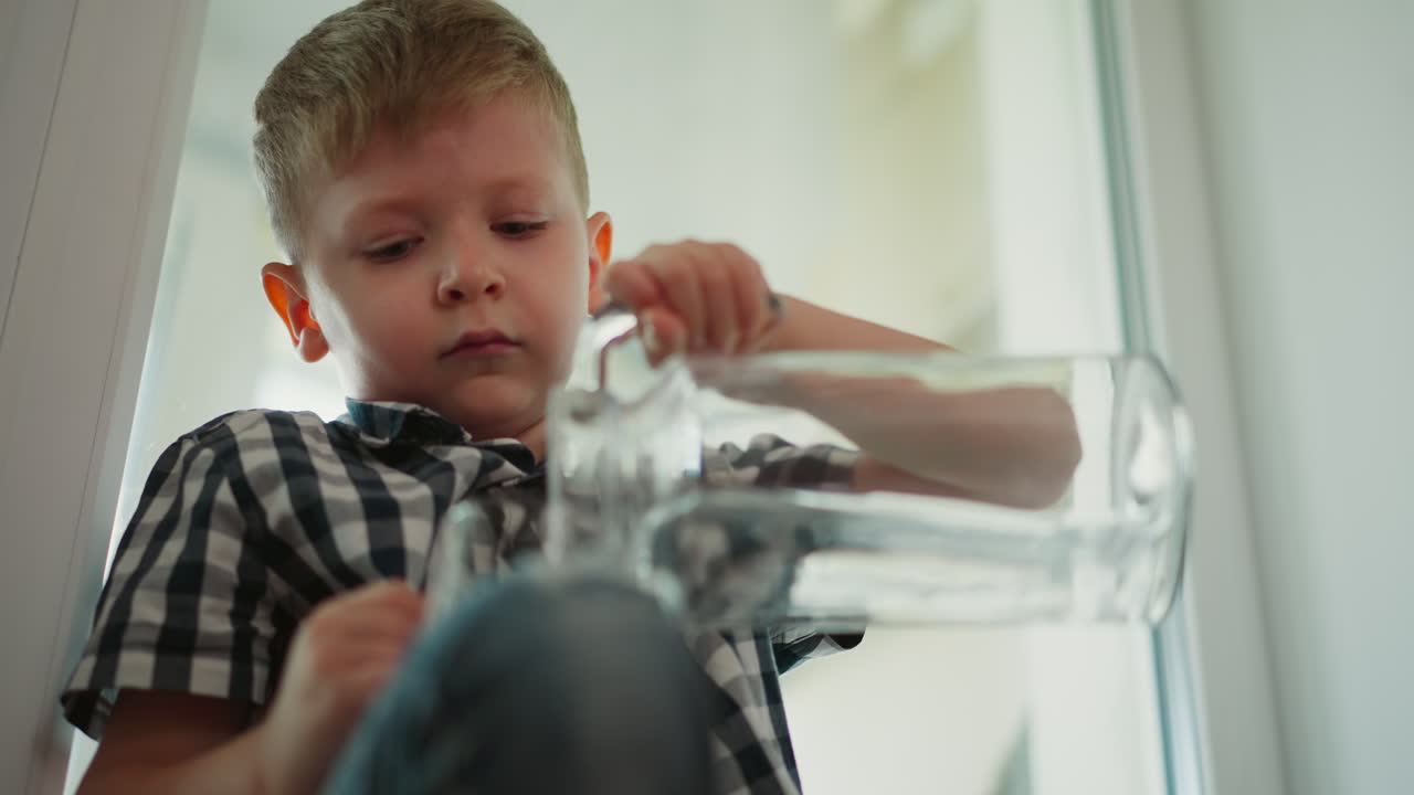 Young boy sitting indoors carefully pours clear water from glass jar into transparent cup wearing plaid shirt focused on task with soft blur background and natural lighting filling room