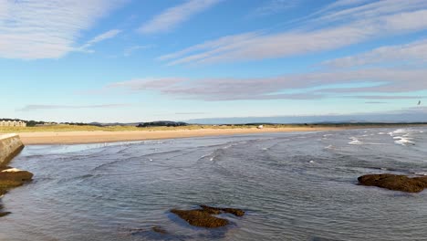 camera pans from a stone bridge to reveal west sands beach, gentle waves, and dramatic skies in bright daylight at st andrews, scotland