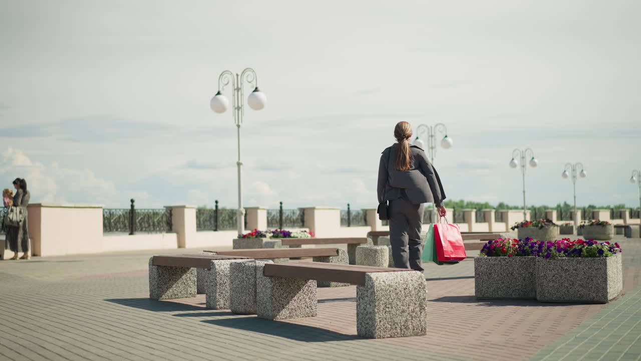 Lady in grey clothing walks towards an outdoor bench, her clothes flutters, she drops her shopping bags, sits down, crosses her legs, and adjusts her hair, two women stand close to the bridge