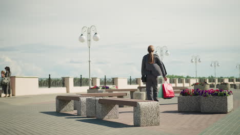 lady in grey clothing walks towards an outdoor bench, her clothes flutters, she drops her shopping bags, sits down, crosses her legs, and adjusts her hair, two women stand close to the bridge