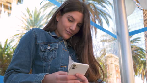 smiling young woman texting on phone sitting at bus stop in a sunny day 1