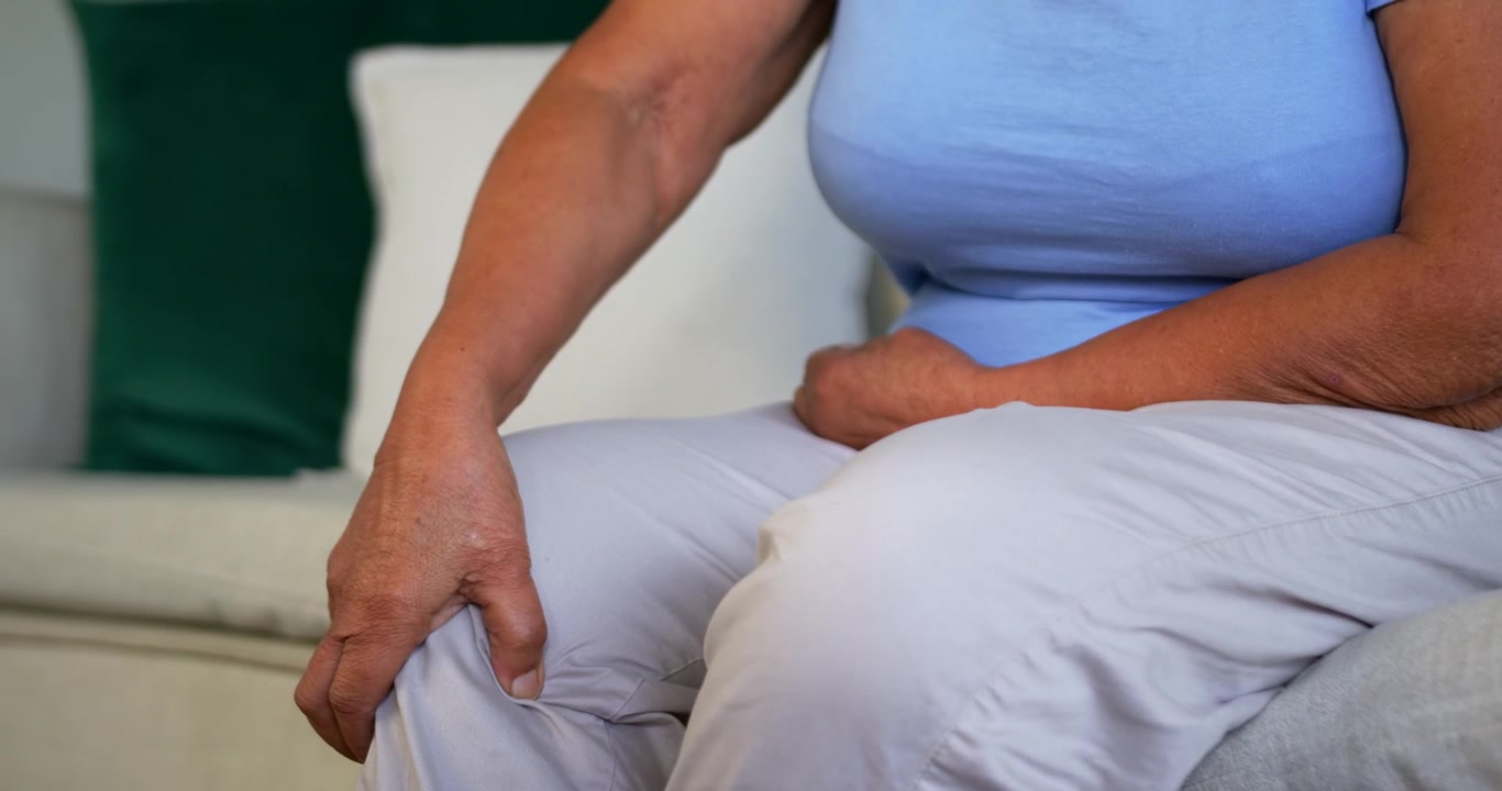 Senior African American female feeling knee pain while rubbing and patting right knee on sofa