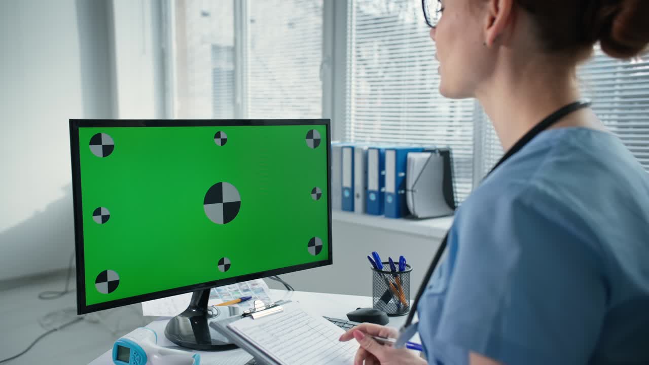 el médico de familia en una cita en línea consulta al paciente a través de una computadora de escritorio con una pantalla verde simulada, la chica en uniforme azul con un estetoscopio escribe en el clipboard