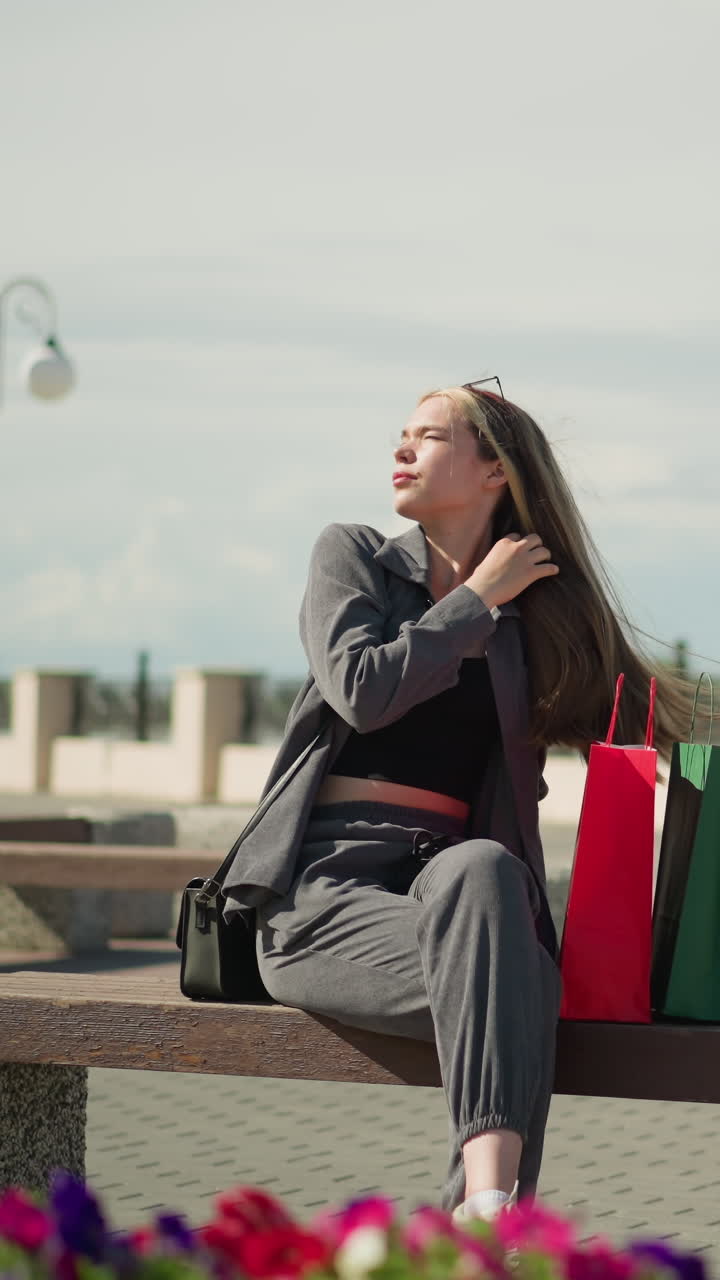 mujer sentada en un banco al aire libre con tres bolsas de compras de colores a su lado, cruza las piernas y ajusta su cabello, mirando hacia arriba en una postura elegante en un día soleado, con un poste eléctrico alrededor