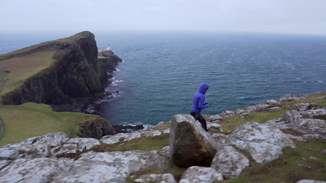 스코틀랜드 스카이 섬에서 사진을 찍으려고 애쓰는 전경과 네이스트 포인트 등대, 대서양을 배경으로 가장자리를 걷고 있는 소녀의 추적 샷