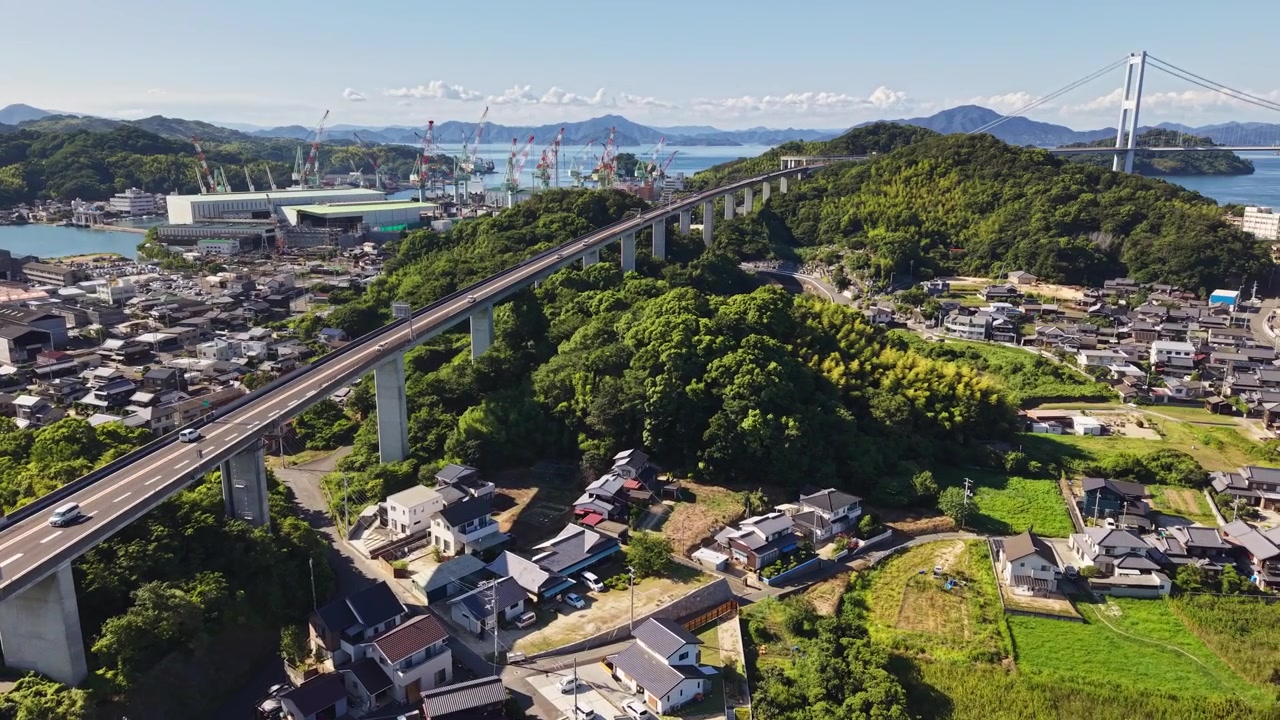 Drone che si allontana sopra un viadotto autostradale sopraelevato, un villaggio costiero, un cantiere navale e un lungo ponte sospeso in Giappone
