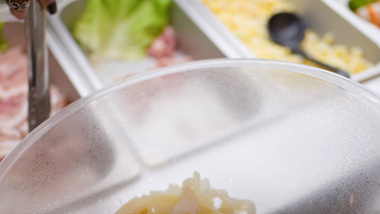 Tongs pick raw sliced meat and vegetables into bowl at bright, self-serve hotpot buffet station