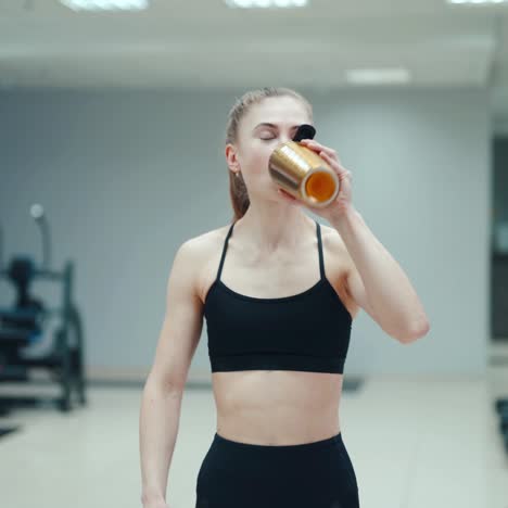 pretty woman in a black top is drinking a drink from a yellow shaker and walking through the gym on the background of sports equipment. healthy lifestyle