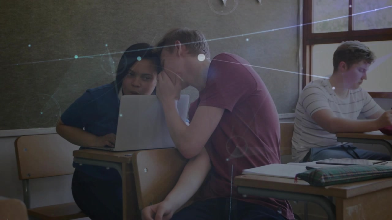 Uomo che sussurra ai compagni che condividono un laptop mentre lavorano a un compito di classe, con bagliori di luce che si sovrappongono alla scena in una scuola.