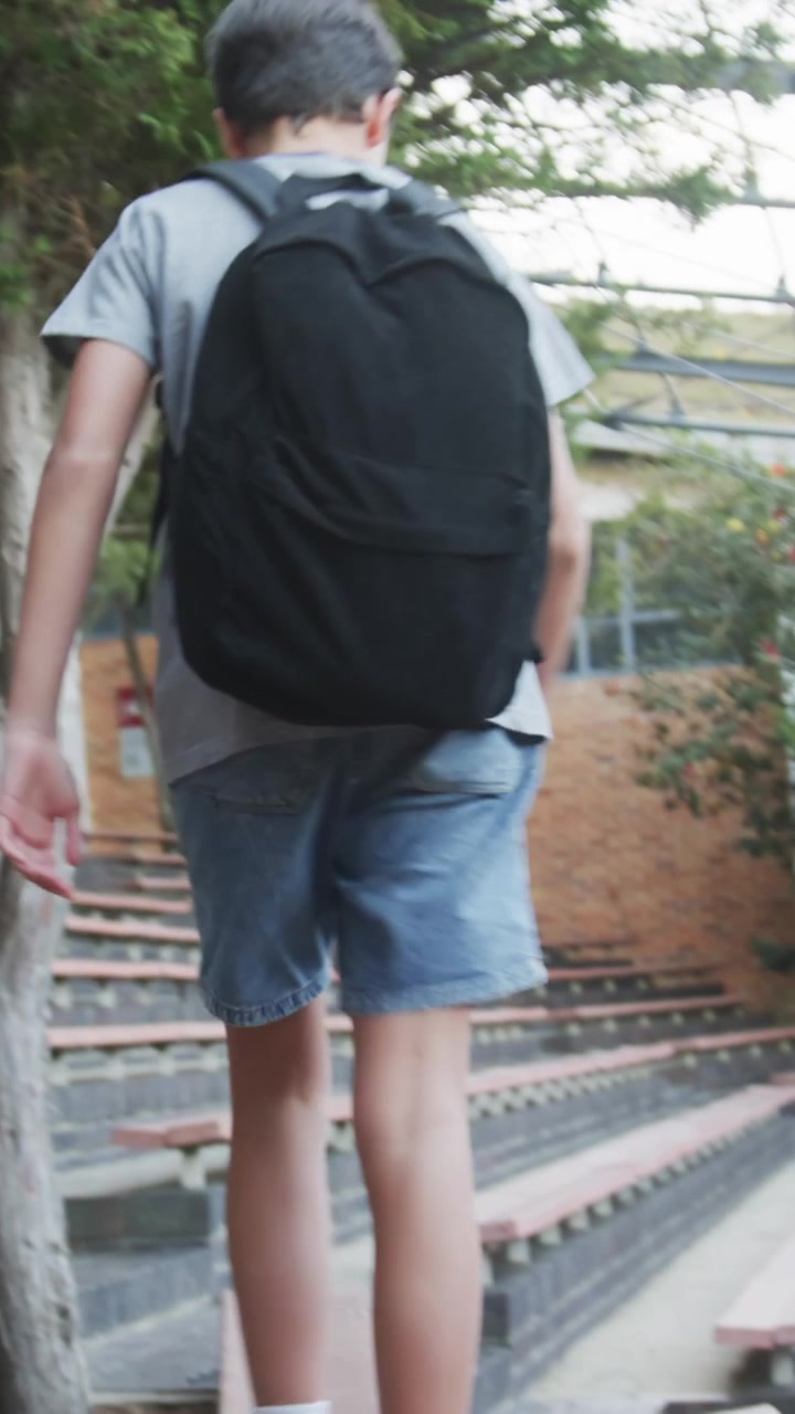Vertical video: Schoolboy turning with backpack, hopping down tiered seats toward door in courtyard