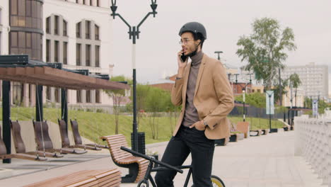 Joven-Americano-Con-Ropa-Formal-Y-Casco-Que-Tiene-Una-Llamada-Por-Teléfono-Móvil-Mientras-Está-Sentado-En-Bicicleta-En-La-Ciudad