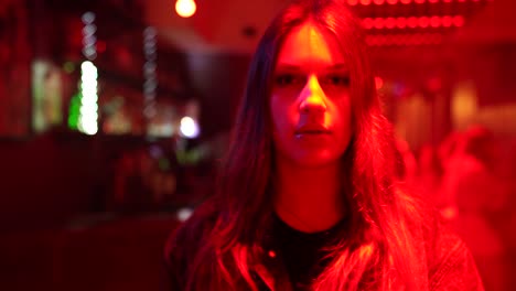 joven mujer caucásica con el pelo largo caminando hacia la cámara, en un bar con iluminación roja
