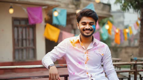 young man is smiling and posing during holi festival celebrations, with colorful gulal powder on his face and clothes, leaning on a railing in a decorated outdoor location