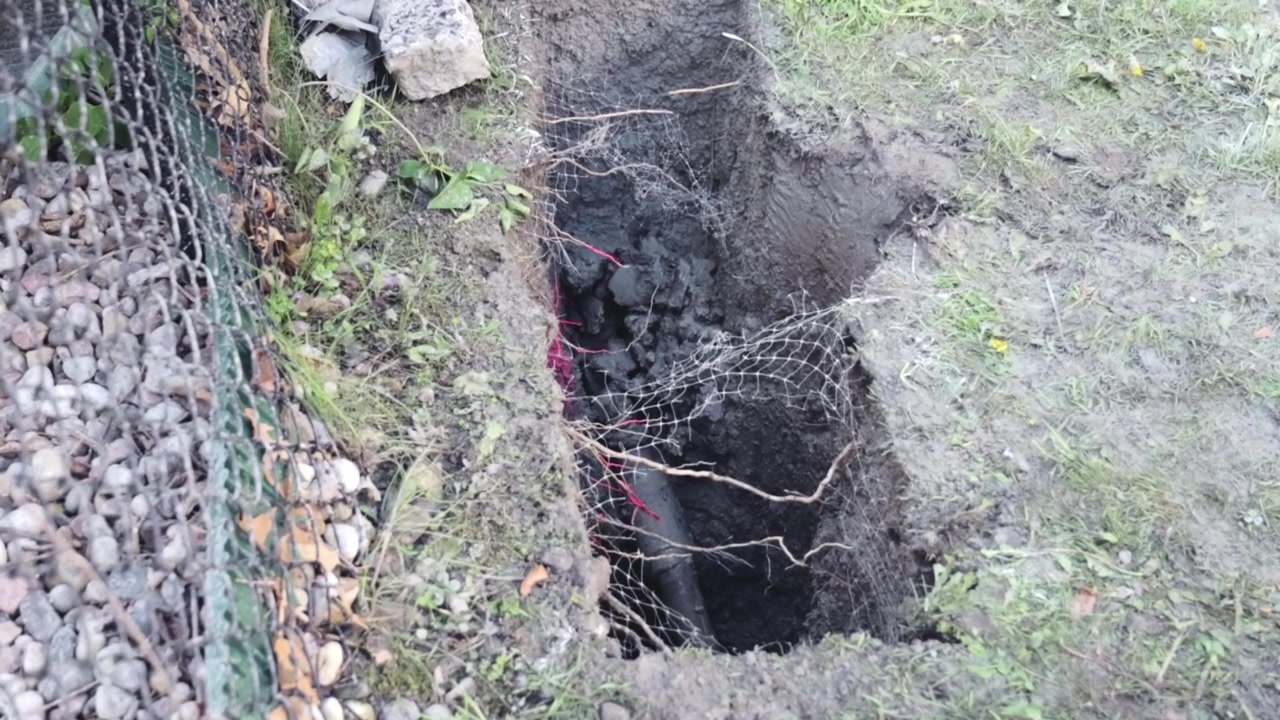A slant‑face drill bit is pushed into an open hydrovac pit where soil and mesh are exposed, guiding electrical cables through the cleared section during daylight.
