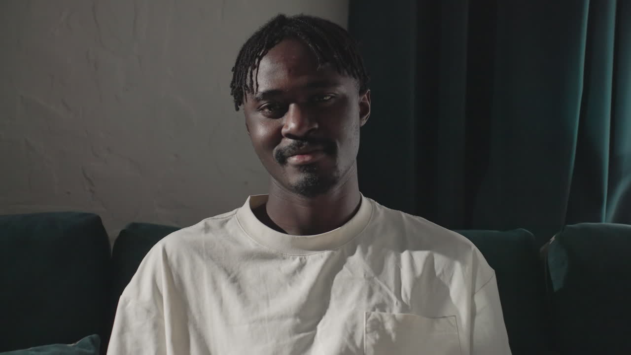 Black youth in white shirt sitting comfortably on couch smiling, looking at camera with calm expression, relaxed in modern home, enjoying leisure time, natural lighting, cozy environment