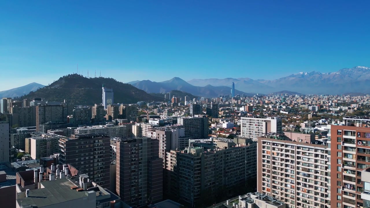 Volo in avanti su un'espansione urbana residenziale con gru edili che rivelano il Cerro San Cristóbal, la torre Costanera Center e la catena delle Ande. Santiago, Cile