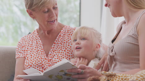 Granddaughter-Sitting-On-Sofa-With-Mother-And-Grandmother-At-Home-Reading-Book-Together