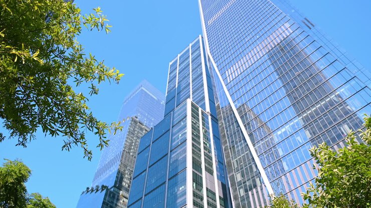 Nova York, EUA, 5 de agosto de 2025: árvores verdes crescem perto dos enormes edifícios modernos. Fachadas de vidro do arranha-céu refletem o céu azul. Visão de baixo ângulo