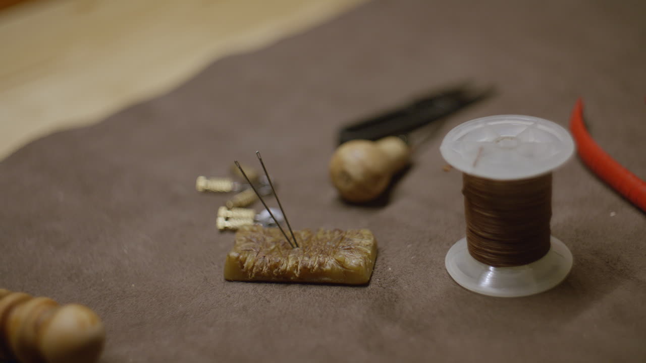 A collection of leather crafting tools, including waxed thread, needles inserted into a beeswax block, an awl, and small metal hardware on a brown fabric surface.