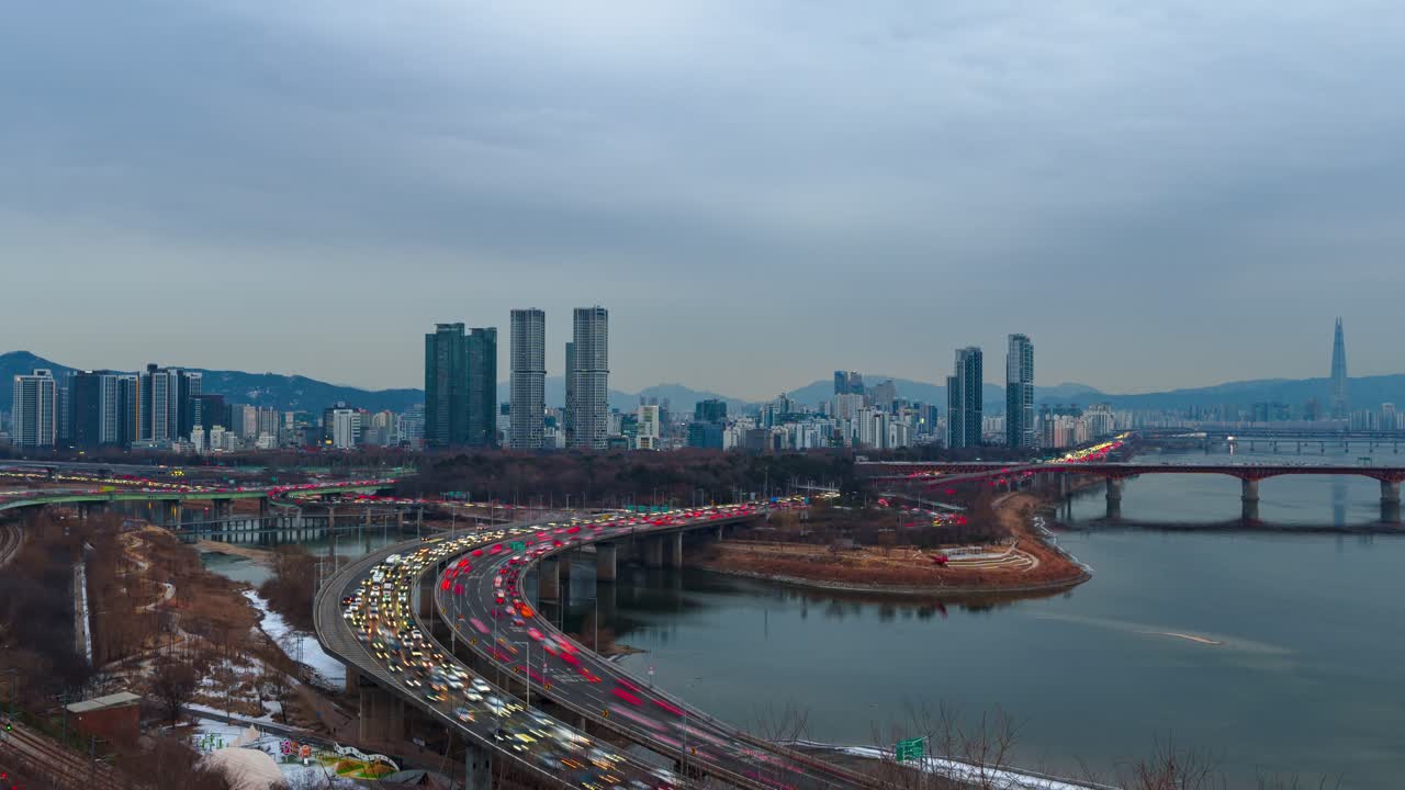 Hyperlapse da giorno a notte di Seoul con zoom indietro, traffico intenso sull'autostrada Gangbyeonbuk-ro, Ponte Seongsu, fiume Han, Lotte Tower, skyline con torri gemelle, montagne, cielo invernale coperto che sfuma nell'ora blu.