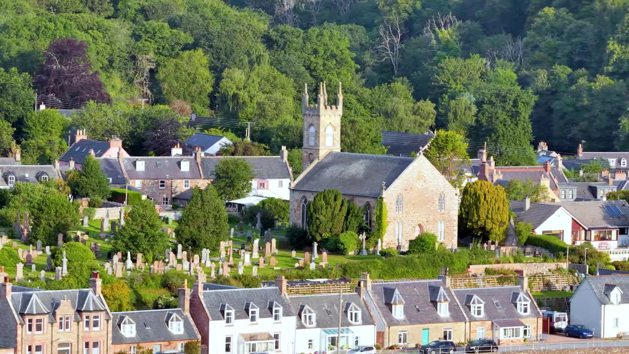 Slow aerial pan reveals stone church, graveyard, village houses, lush greenery, and evening sunlight