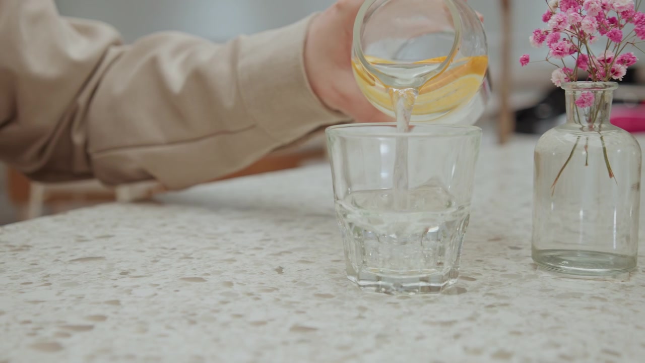 Primo piano di una mano che versa acqua con fette di limone in un bicchiere su un piano di lavoro maculato, accanto a un piccolo vaso con fiori rosa. La scena presenta un'illuminazione soffusa e un movimento minimo della fotocamera, focalizzando l'attenzione.