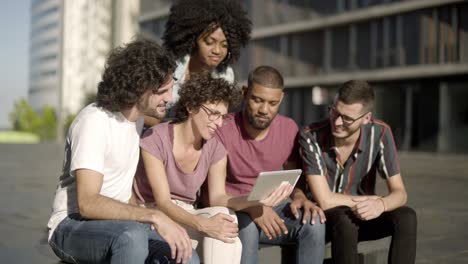 Cheerful-friends-sitting-on-wooden-bench-and-looking-at-tablet