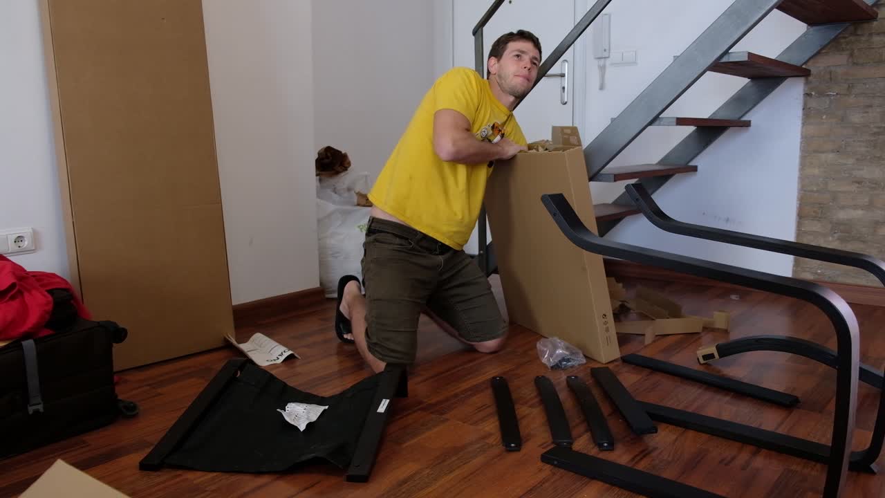 젊은 남자가 마분지 상자에서 조립되지 않은 가구 조각을 풀고 있습니다.