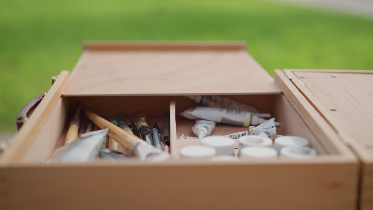 artist sets up paintbox in park, places palette on bench and arranges brushes and tubes hands reach into wooden case, sunlight on grass, casual plein air sketching session with relaxed mood