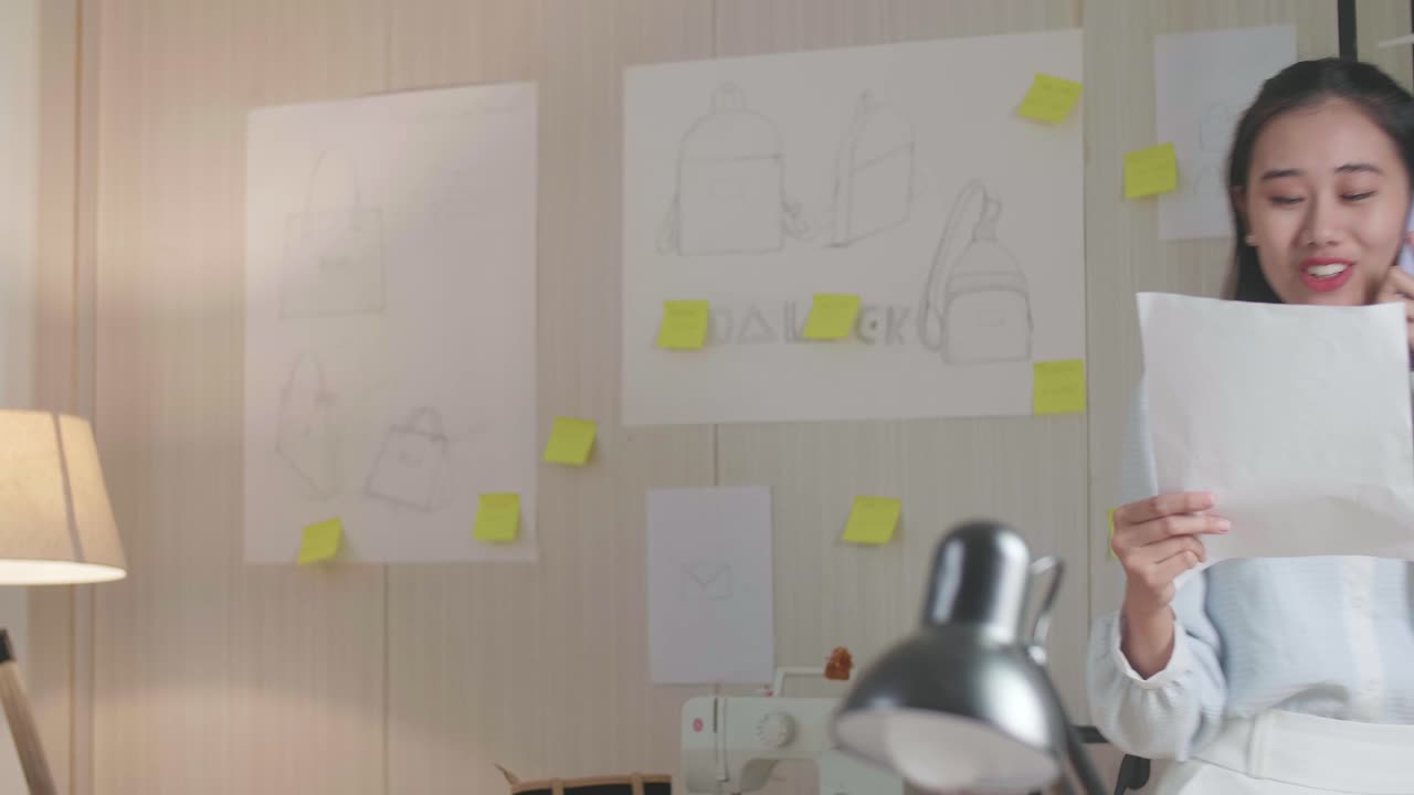 diseñadora asiática hablando en un teléfono inteligente mientras mira el enlace de diseño en la mano y caminando en la oficina