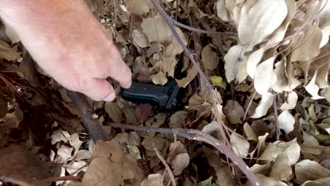 man pulling out a black firearm stashed hidden in some bushes and dry leaves