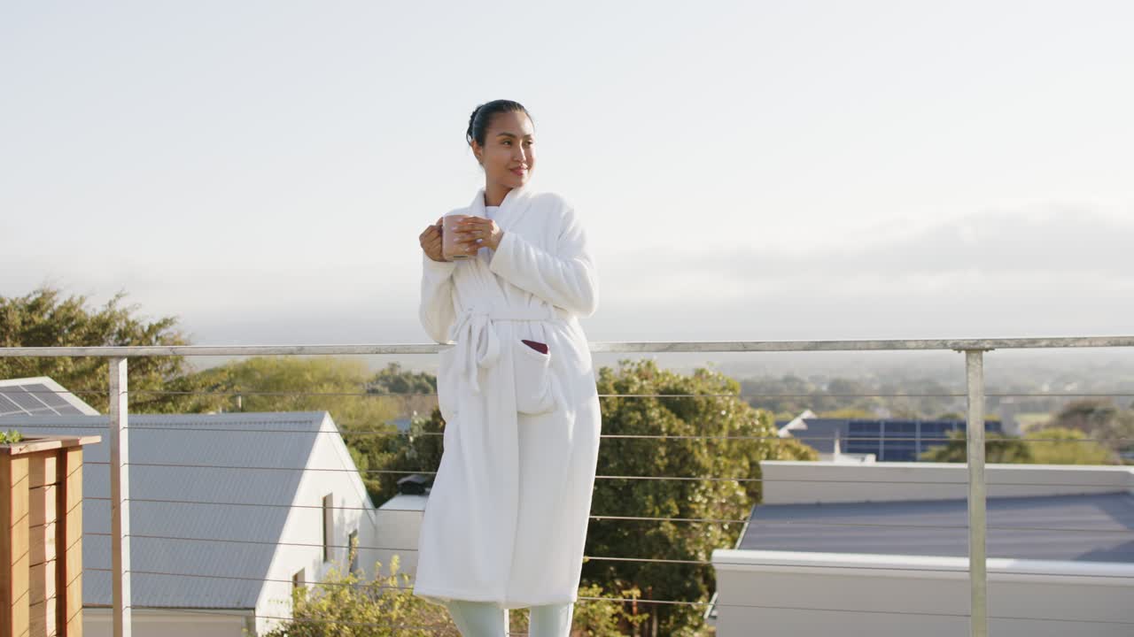 Morning sun inspiring mid adult Asian woman sipping from ceramic mug on terrace and gazing rooftops