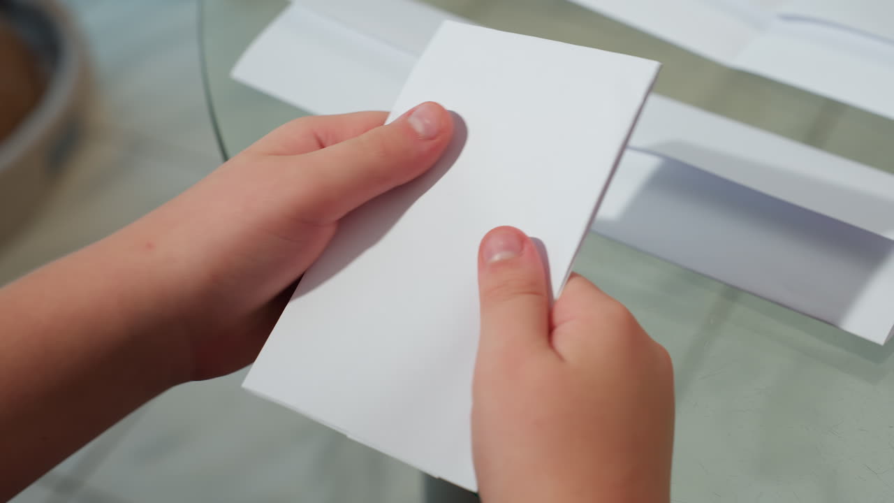 Hand view of kid folding paper on table, focused on creating something, white paper visible, showcasing a hands-on creative activity in a home setting